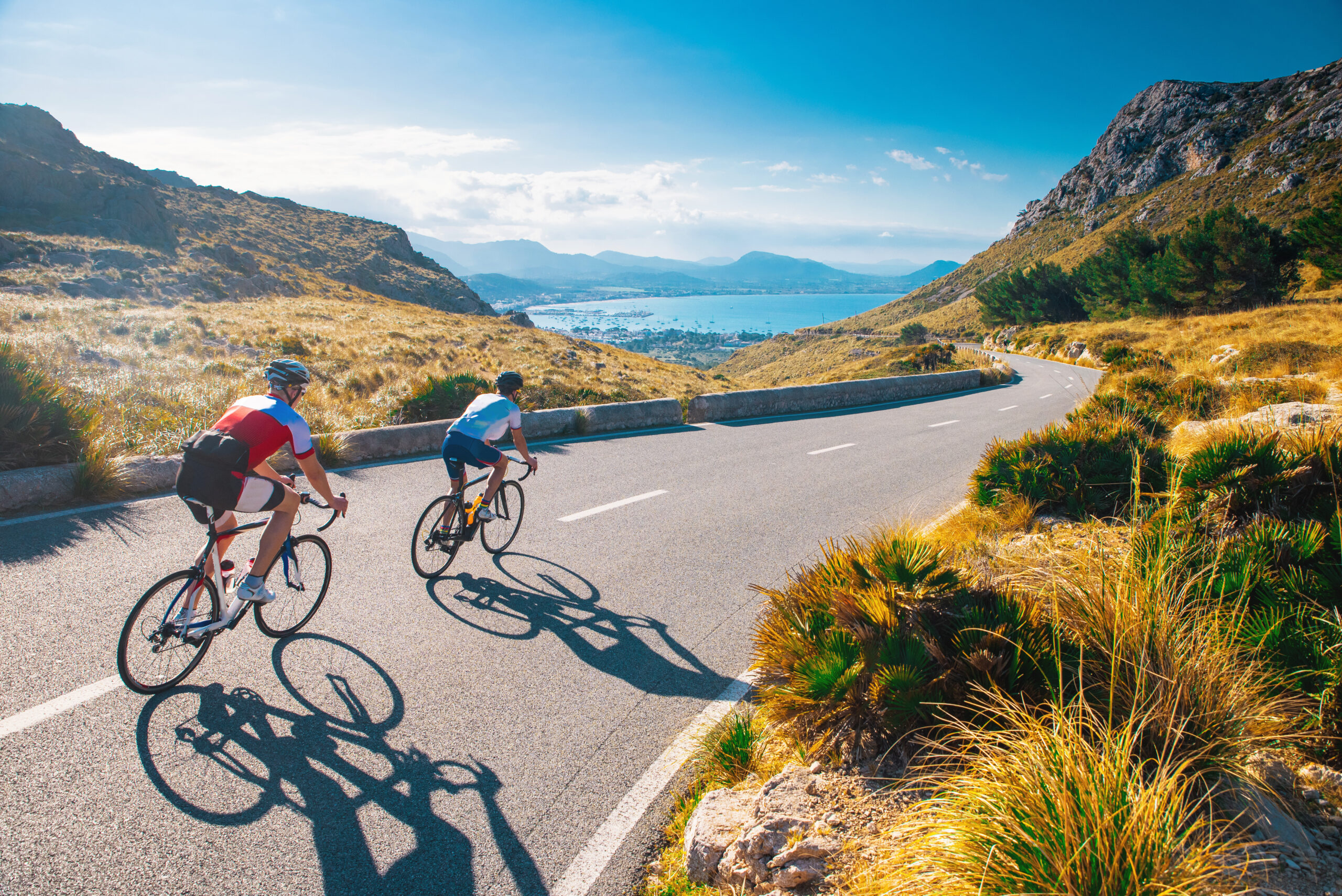 Two people cycling down a winding road in Alcudia with the sea and mountains in background.